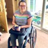 Image is a photograph of writer and wheelchair user Elizabeth Gwilliam using the Trabasack Mini on her lap as a wheelchair tray to hold a hot drink and a remote control