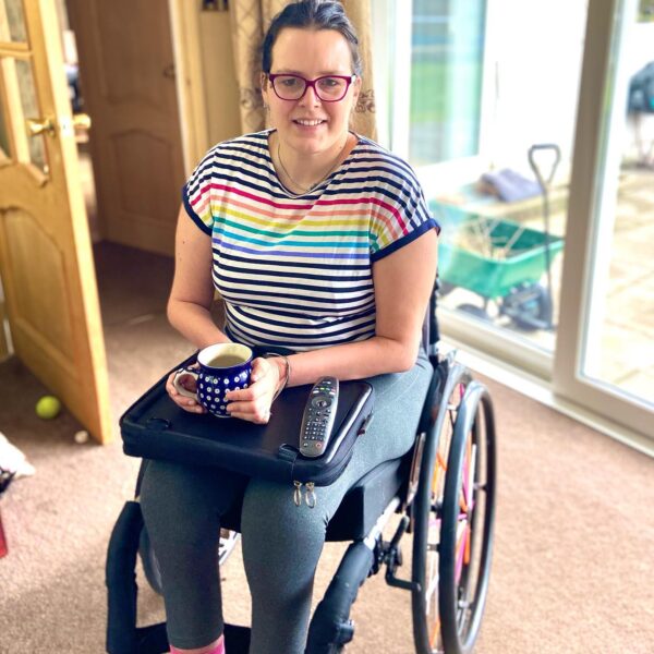 Image is a photograph of writer and wheelchair user Elizabeth Gwilliam using the Trabasack Mini on her lap as a wheelchair tray to hold a hot drink and a remote control