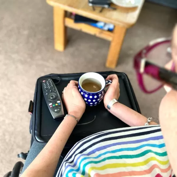 Image is a photograph viewing the Trabasack Mini wheelchair tray and bag in one on the lap of Elizabeth, from her visual perspective. On the Trabasack Mini tray is a mug of hot drink and a remote control