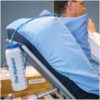A man reclines on a hospital-style bed with a Hydrant water bottle hanging from the side rail. A drinking tube extends from the bottle to near his mouth.