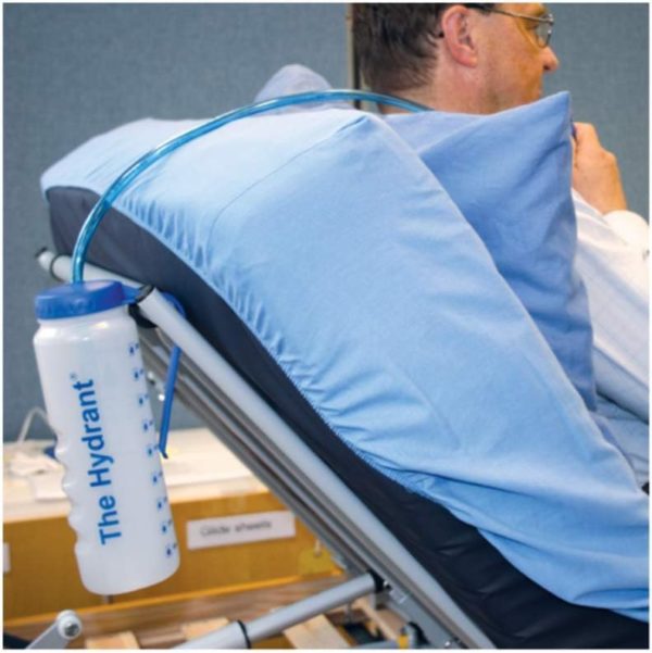 A man reclines on a hospital-style bed with a Hydrant water bottle hanging from the side rail. A drinking tube extends from the bottle to near his mouth.