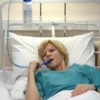 A woman in hospital clothing lies in bed and drinks from a blue bite valve connected to a Hydrant water bottle hanging from the bed frame.