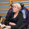 A seated woman in a wheelchair sips from a tube connected to a Hydrant water bottle clipped to her chair. She wears glasses and a black top.