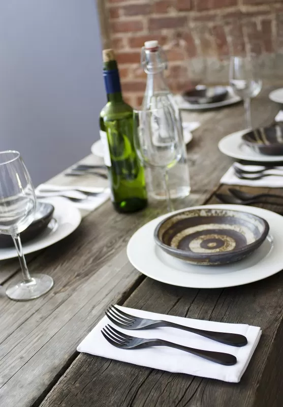 Image is a photograph of a stylish weathered-wood dining table laid with wine glasses, earthenware pottery and matt black finish Knork cutlery