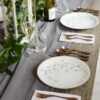 Image is a photograph of a wooden table laid with pretty floral decoration, a bottle of wine, plates and Knork cutlery in an Antique Brass finish