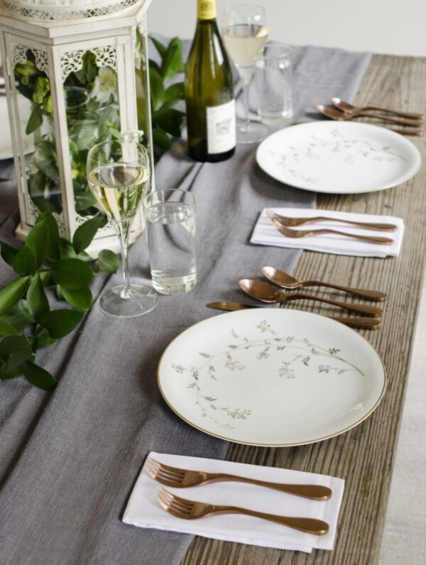 Image is a photograph of a wooden table laid with pretty floral decoration, a bottle of wine, plates and Knork cutlery in an Antique Brass finish
