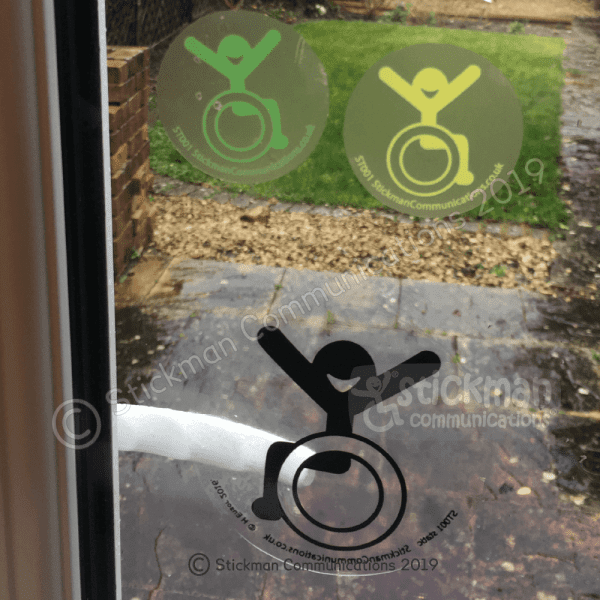 Image is a photograph of three Happy Accessibility stickers in green, yellow and black on a glass window