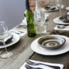 Image is a photograph of stylishly-weathered wooden table in an alfresco setting, laid with earthenware plate, wine bottles and glasses, and Knork cutlery in a Matt Black finish