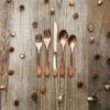 Image is a photograph of a rustic, wooden table scattered with pinecones and acorns, with a set of Knork cutlery in an Antique Brass finish set centrally on the table