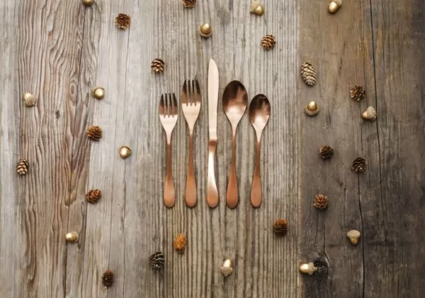 Image is a photograph of a rustic, wooden table scattered with pinecones and acorns, with a set of Knork cutlery in an Antique Brass finish set centrally on the table