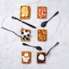 Image is a photograph of 6 square plates of food on a marble surface with Matt Black Knork cutlery