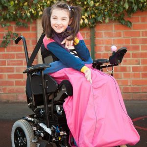 Image is a photograph of a young girl with brown hair in bunches, sat smiling in a wheelchair with a pink waterproof leg cover