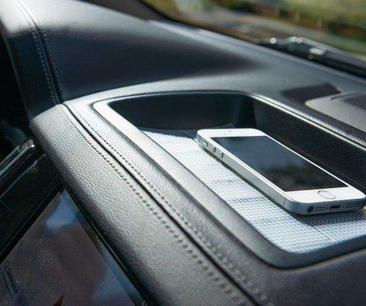 Image is a photograph of a mobile phone sitting atop a strip of Cat Tongue Grip Tape on a car dashboard