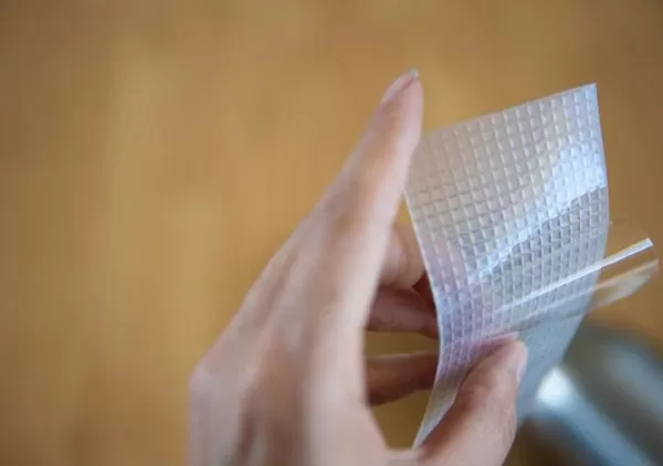 Image is a photograph of a woman's hand peeling-back the protective film on the adhesive side of a strip of Cat Tongue Grip tape