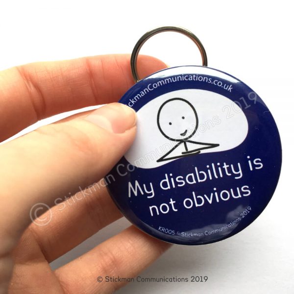 Image is a photograph of a hand holding a blue, round keyring with a smiling stickman illustration, with text which reads: "My disability is not obvious"