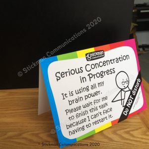 Image is a photograph of a sign with a rainbow-coloured border, with a an illustration of a stickman wearing reading glasses, looking intently at a piece of paper. Text reads: "Serious concentration in progress. It is using all my brain power. Please wait for me to finish this task because I can't face having to restart it. DO NOT DISTURB"