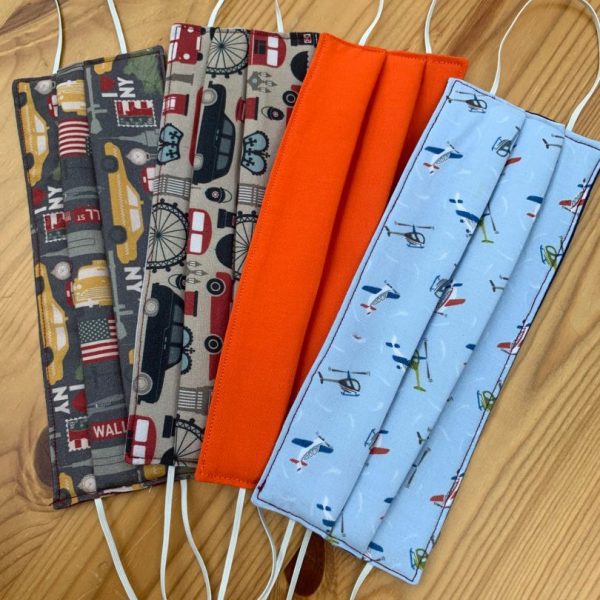 Image is a photograph of four fabric face masks, laying flat on a wooden table top. Each mask has a different design, including New York, London, plain orange and Aviation