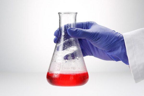 Image is a photograph of an a hand in a nitrile glove holding out a wide-neck conical lab flask, partially filled with red liquid.