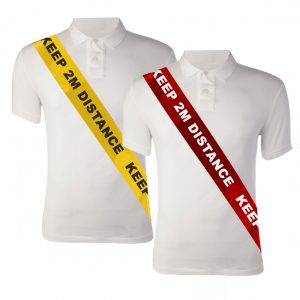 Image is a photograph of two mannequin torsos wearing white polo shirts. Each mannequin wears a silky social distancing sash from the shoulder, around the chest and to the waist. The first sash is yellow with black text, the second is red with white text. Capitalised text reads "KEEP 2M DISTANCE"
