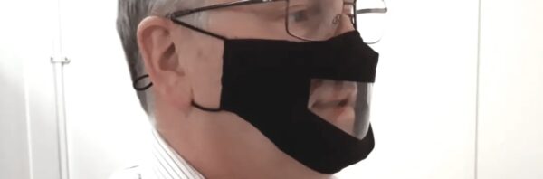 Image is a photograph of the profile of an older man in glasses, wearing a black lip reading mask with transparent panel over the mouth and nose area