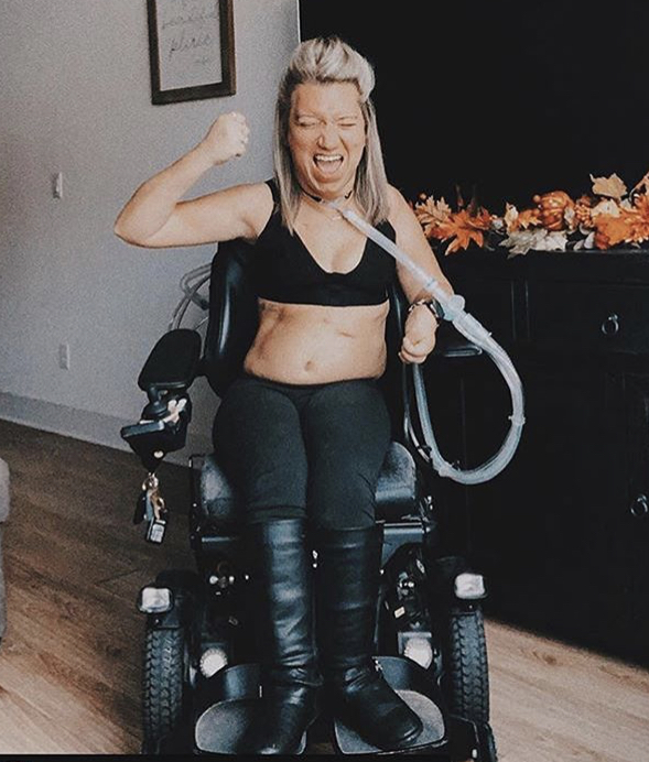 Image is a photograph of a lady with blonde hair, wearing black knee-high boots, leggings and a black Elba London bra, sat in a powerchair, triumphanty raising a clenched fist upwards with a closed-eyed, cheerful expression on her face.