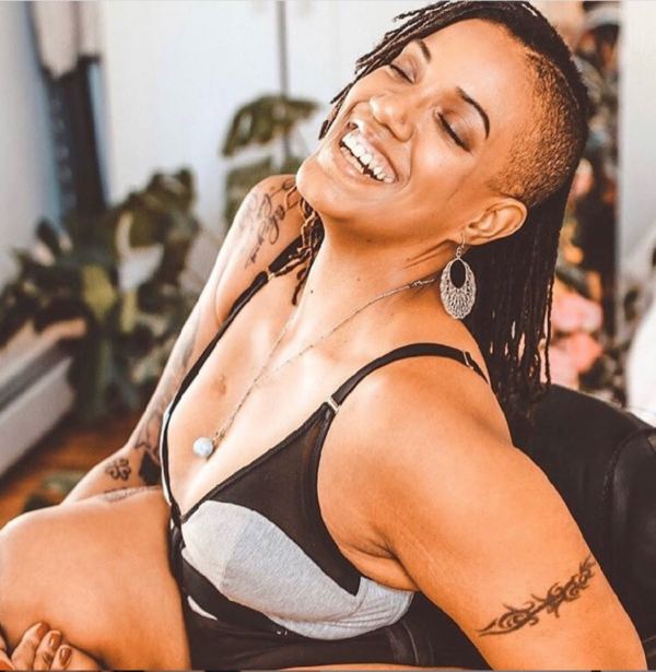 Image is a photograph of Krystal smiling, eyes-closed and head tilted backwards, whilst sitting in a chair wearing an Earl grey and black mesh Elba London bra