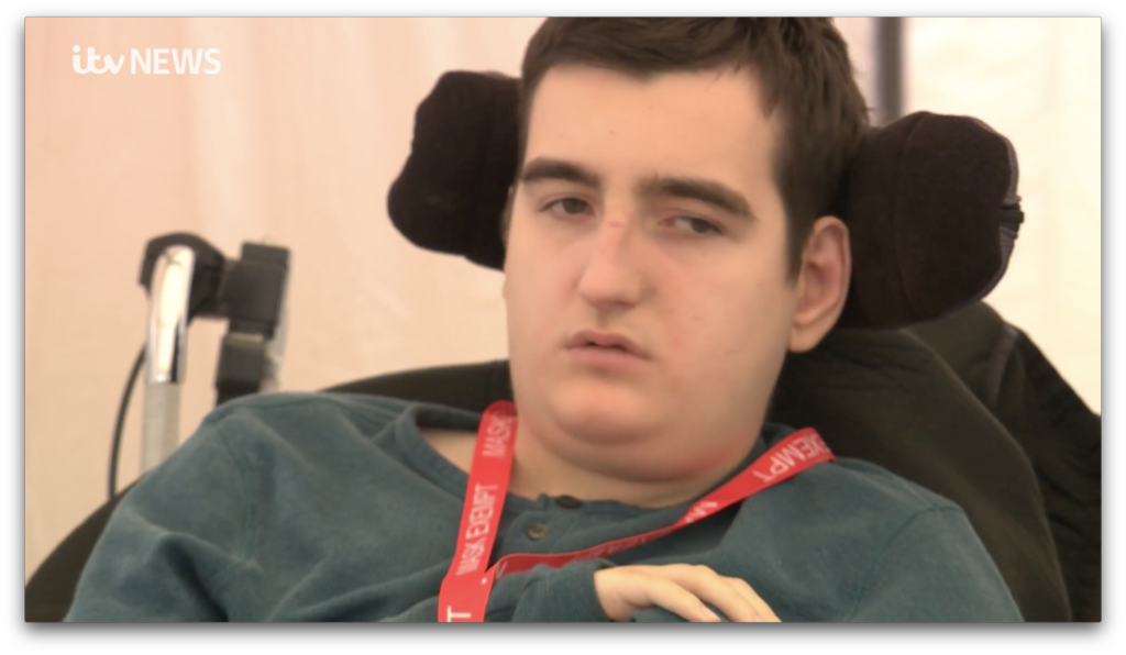 Image is a still from the ITV news segment about Mask Exemption ID cards. The image shows Joe Edwards in a wheelchair, wearing a red lanyard with white text which reads "MASK EXEMPT"
