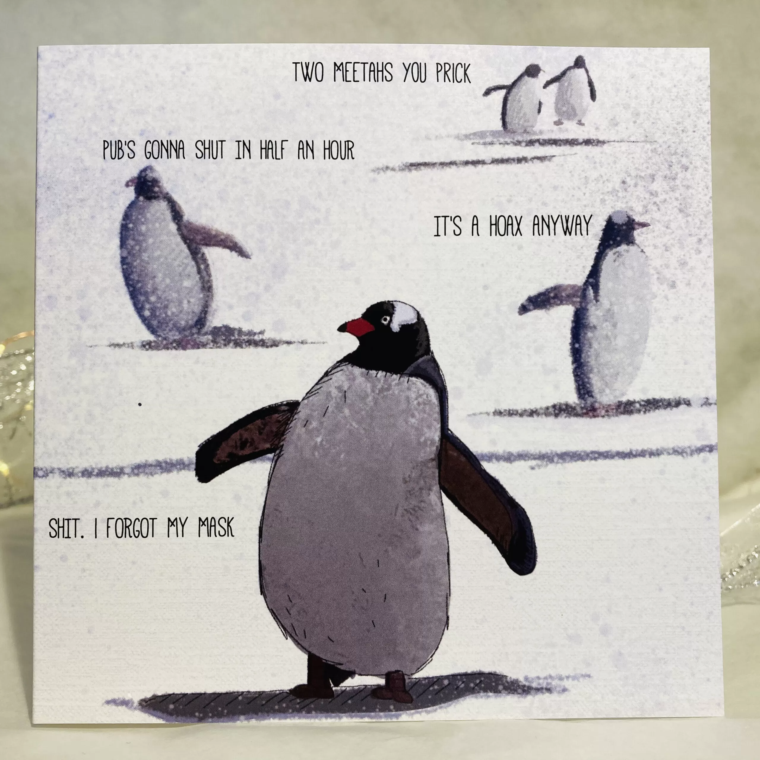 Image is a photograph of an illustrated christmas greeting card featuring penguins walking through the snow. Each penguin is making a humorous statement about Covid-19. Text reads: "TWO MEETAS YOU PRICK. PUB'S GONNA SHUT IN HALF AN HOUR. IT'S A HOAX ANYWAY. SHIT, I FORGOT MY MASK"