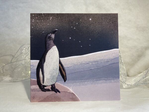 Image is a photograph of a penguin art christmas card, with an illustration of a penguin in the Antarctic, wistfully gazing towards the star-strewn night sky.