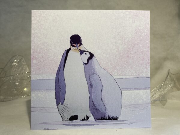 Image is a photograph of an illustrated card featuring a baby penguin looking up to their adult penguin.