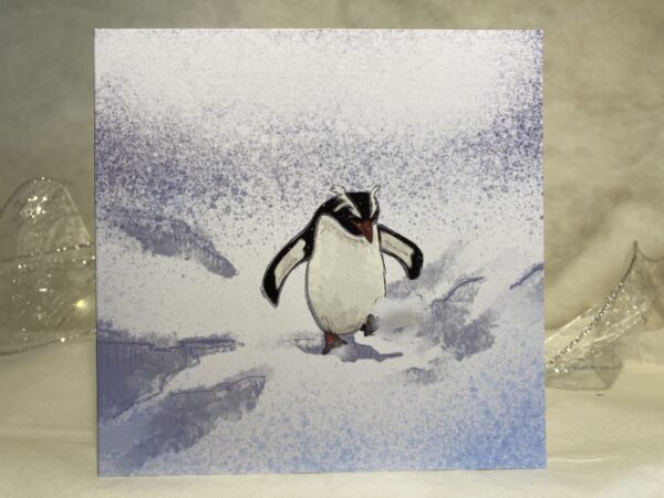 Image is a photograph of an illustrated christmas card featuring a penguin playfully kicking through the snow.