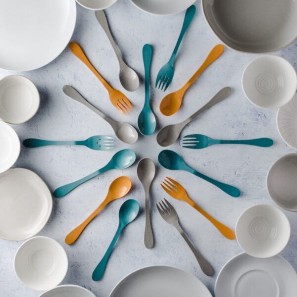 Image is a photograph of Knork Eco cutlery in teal, grey and orange, arranged in a circular pattern surrounded bowls and plates