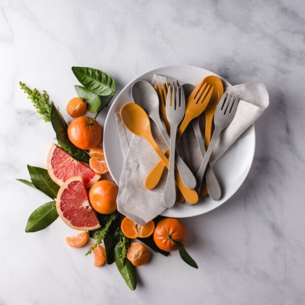 Image is a photograph of a white side plate with orange and grey Knork Eco cutlery on top. The plate is on a grey, marble worktop with a carefully arranged selection of citrus fruits to one side