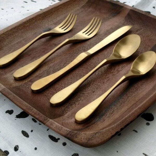 Image is a photograph of a set of Knork cutlery in a light gold, Satin Brass on a dark wood plate with curved corners