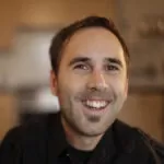 Image is a photographic portrait of Martyn Sibley, smiling directly at the camera whilst wearing a black shirt with epaulets.