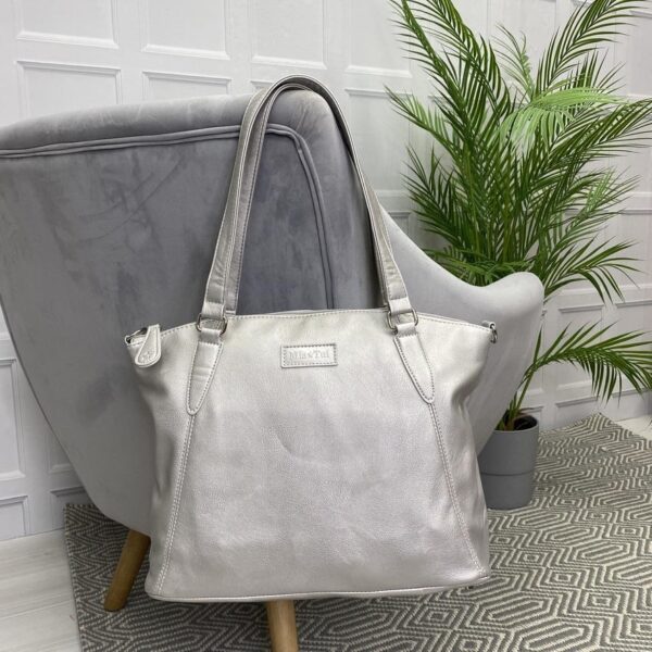 Image is a photograph of a Samantha Renke accessible handbag in Matt Silver hung over the back of a grey velvet armchair in a modern living room