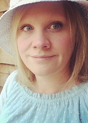 Image is a photograph of Amrick Ainley Disability Horizons shop tech and design manager, wearing a pale green top and white sun hat, smiling whilst sat outdoors.