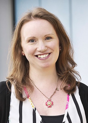 Image is a photograph of Liz Ransome-Croker wearing a black and white top and floral necklace, smiling widely directly to the camera