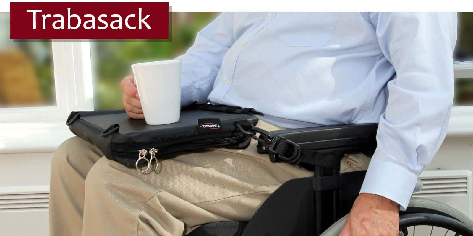 Image is a photograph of a man sat in a wheelchair with a Trabasack Mini lap tray bag on his lap, using the built-in tray to carry a mug of drink. In the top lefthand corner of the image text reads 