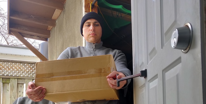 Tony Awad inventor of the T-pull, closing a door whilst carrying a box in a wheelchair, Tony has a beanie hat and is on the porch of his Canada home