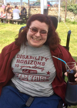 Image is a photograph of Emma Purcell sat outdoors in a beer garden, smiling with a drink in hand and wearing a Disability Horizons t-shirt