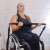 A woman in a wheelchair exercises with a resistance band in a home setting. She is wearing a black tank top and leggings, and she has long hair tied back. A beige sofa and a black TV stand with books are in the background.