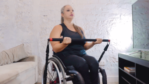 A woman in a wheelchair exercises with a resistance band in a home setting. She is wearing a black tank top and leggings, and she has long hair tied back. A beige sofa and a black TV stand with books are in the background.