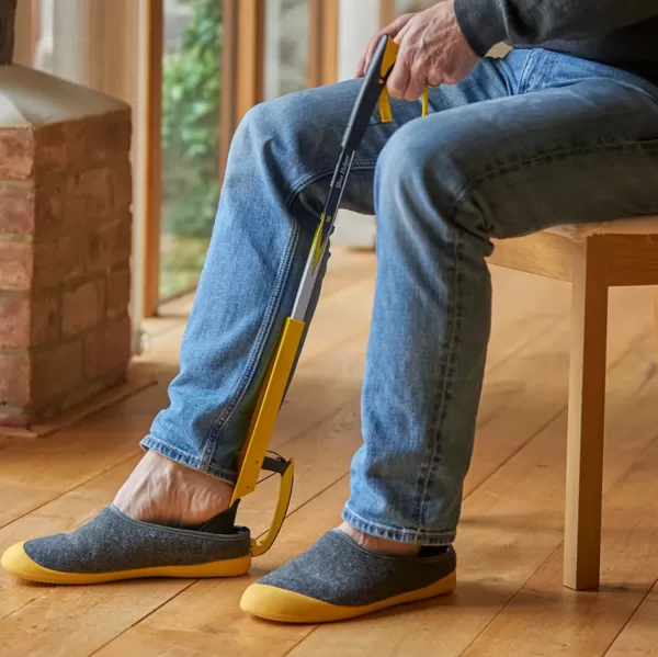 Shoe reacher grabbing aid stick with a shoe horn, a man sits on a chair and uses the yellow aid to help him but on shoes