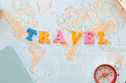Travel image showing the words travel spelt out in yellow, blue and pink letters on a map of the world with a compass and notebooks