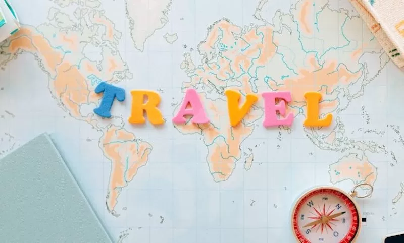 Travel image showing the words travel spelt out in yellow, blue and pink letters on a map of the world with a compass and notebooks