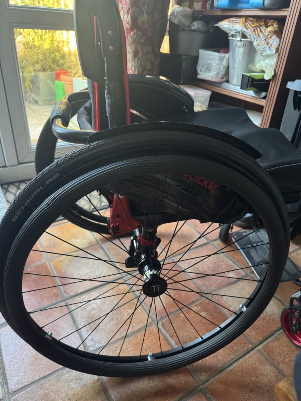 A close-up of a wheelchair's rear wheel, showing a black Schwalbe tire with a titanium hub and metal spokes, rehadesign groovy pushrim covers are on the pushrims. The frame is red with black accents, and a curved push handle is visible. The background includes a tiled floor, a shelving unit with household items, and a glass door leading outside.