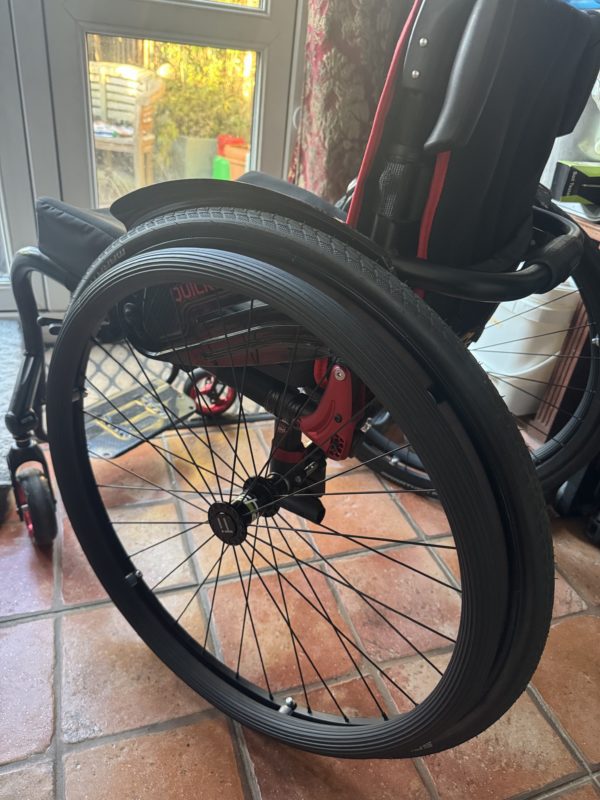 A close-up of a wheelchair’s rear wheel, featuring a black Schwalbe tire with a titanium hub and metal spokes. The pushrim is covered with RehaDesign Groovy textured covers for improved grip. The frame is red with black accents, and a curved push handle is visible. The background includes a tiled floor, a glass door leading outside, and part of a shelving unit with household items.