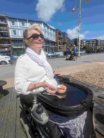 Clare using a trabassack to eat seafood on a seawall, she is a power chair and there are hotels in the background and the stoney beach of bognor regis can be seen