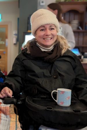 Lady with a white hat and anorack in a power chair, smiling with a trabasack curve wheelchair tray with a cup on it
